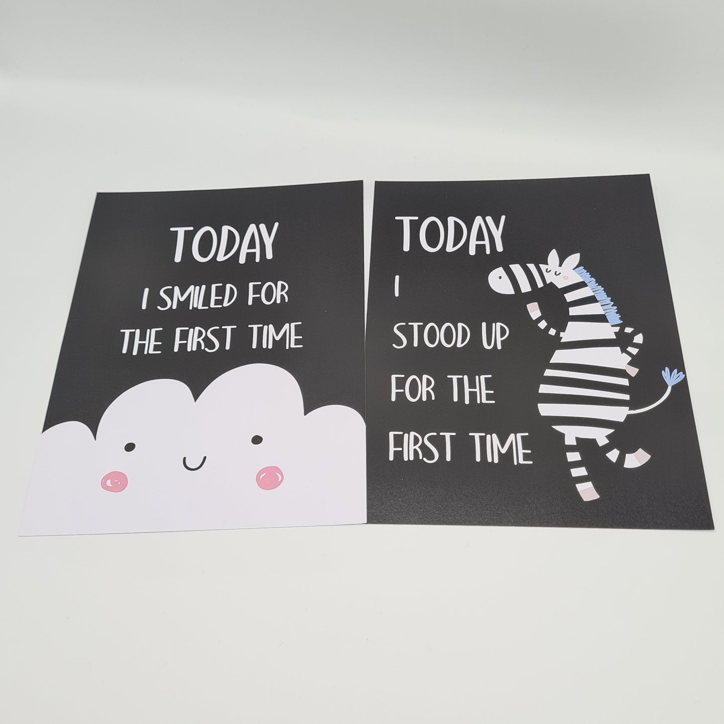 Two baby milestone cards, both black in colour. The card on the left features an illustration of a white cloud at the bottom with white text above saying "Today I smiled for the first time". The card on the right features an illustration of a standing zebra with white text "Today I stood up for the first time".