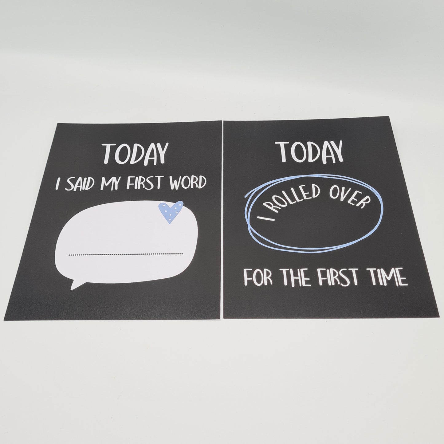 Two baby milestone cards, both black in colour. The card on the left features a white speech bubble with a little blue heart and a dotted line for something to be written on. Above it there is white text saying "Today I said my first word". The card on the right contains the text "Today I rolled over for the first time".