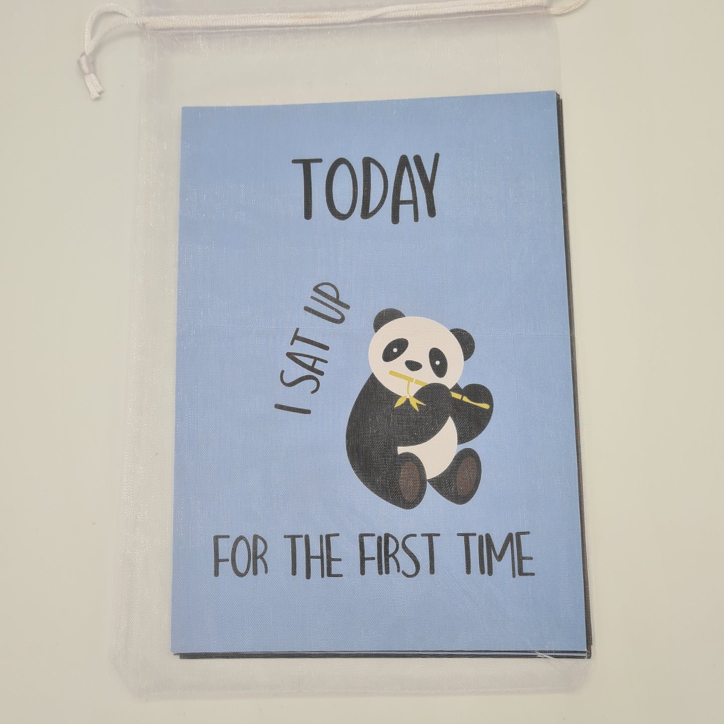 Pack of blue and black Baby Milestone cards encased in a white organza bag. The visible top card is blue and contains an illustration of a panda sitting up and eating bamboo with the text "Today I sat up for the first time".