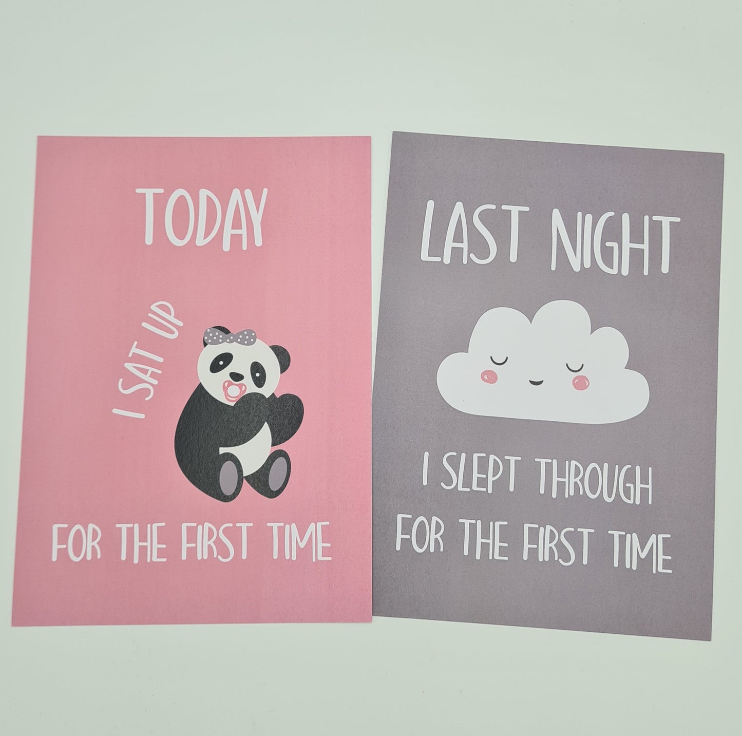 Two baby milestone cards. The card on the left is pink and features an illustration of a sitting baby panda with the text "Today I sat up for the first time". The card on the right is grey and features an illustration of a white cloud with a face and the text "Last night I slept through for the first time".