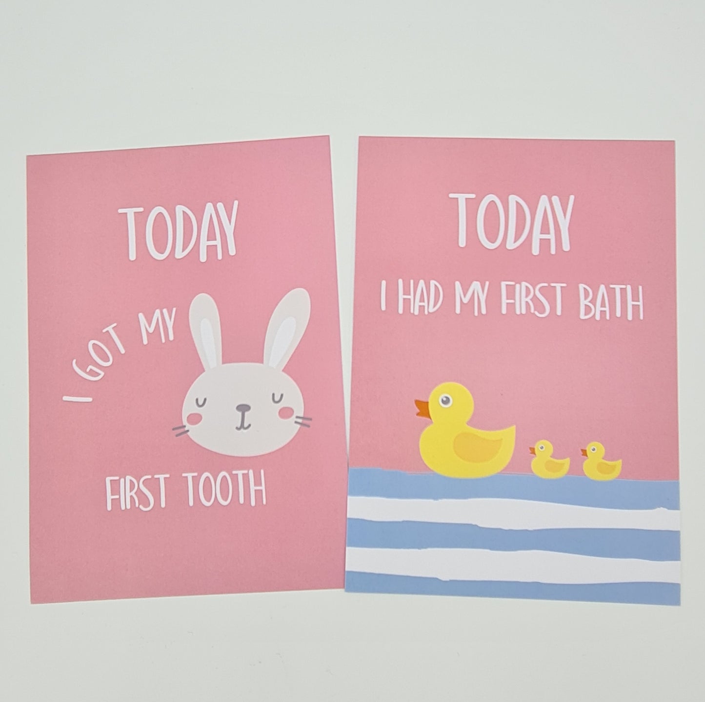 Two pink baby milestone cards. The card on the left features an illustration of a rabbit's face and the text "Today I got my first tooth". The card on the right features an illustration of three yellow ducks on blue and white water and the text "Today I had my first bath".