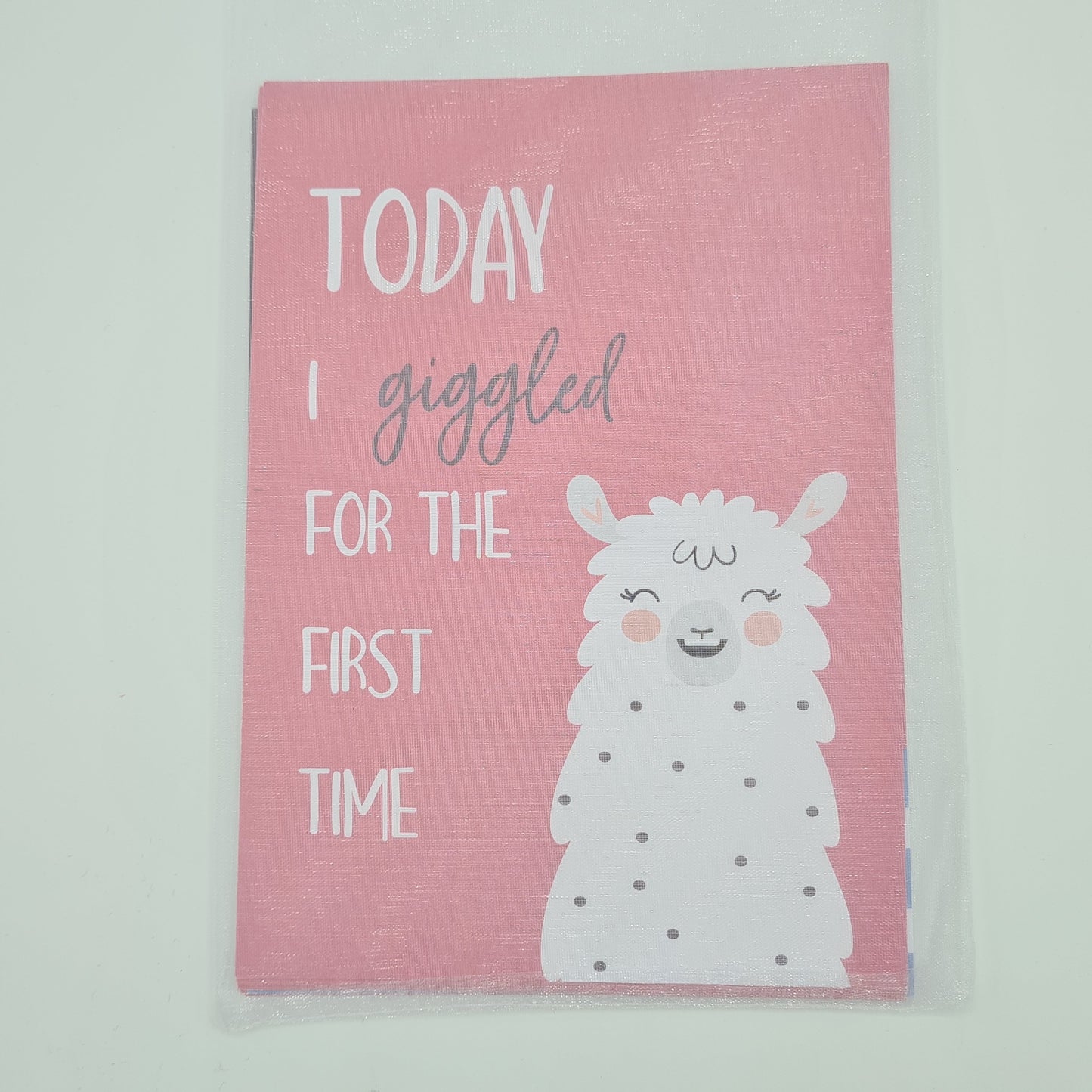 Pack of pink Baby Milestone cards encased in a white organza bag. The visible top card contains the text "Today I giggled for the first time" alongside an illustration of a laughing llama.