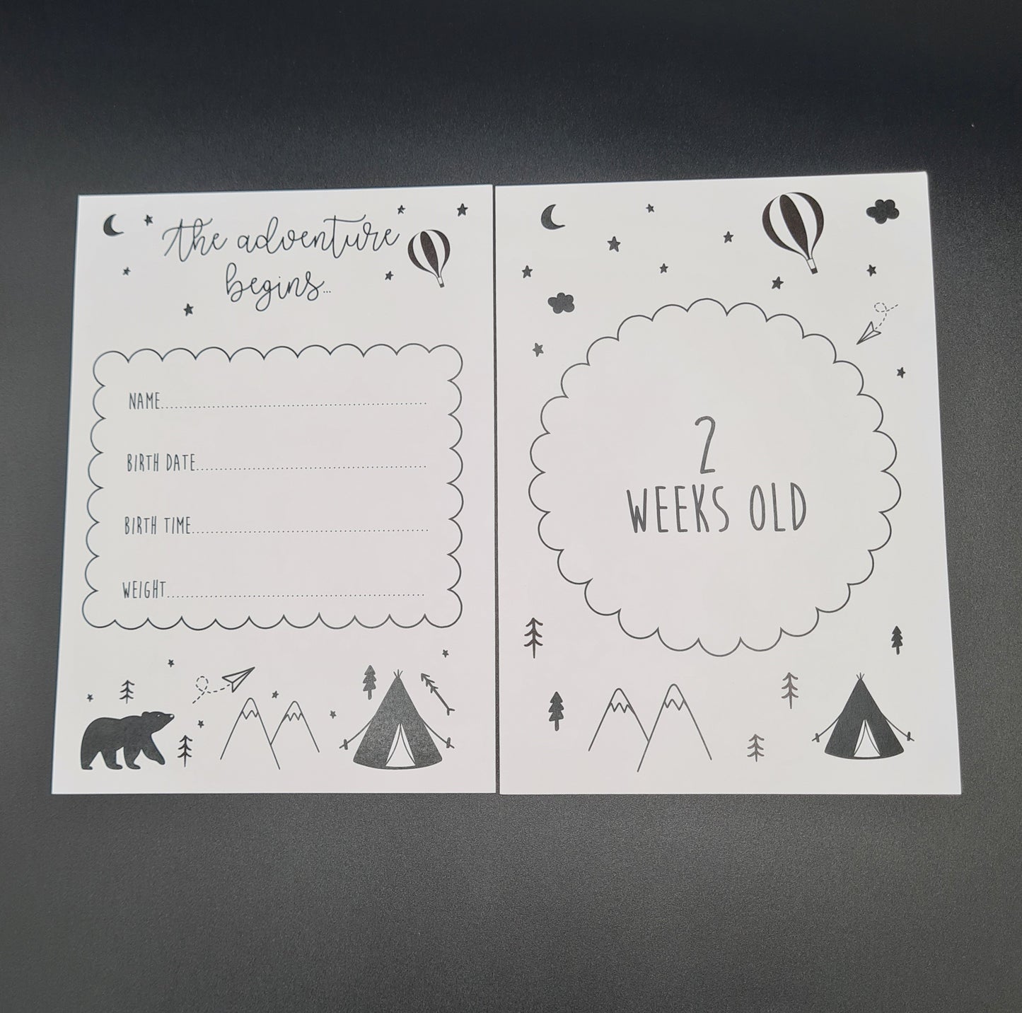 Two white baby milestone cards. At the top of the card on the left is the text "The adventure begins" with an illustration of a hot air balloon, the moon and some stars. There is a box in the middle of the page with Name, Birth Date, Birth Time and Weight titles next to dotted lines upon which the information can be written. Below that is an illustration of a tent, some mountains and a bear. The card on the right contains similar illustrations and has the text "2 Weeks Old" contained within a circle.