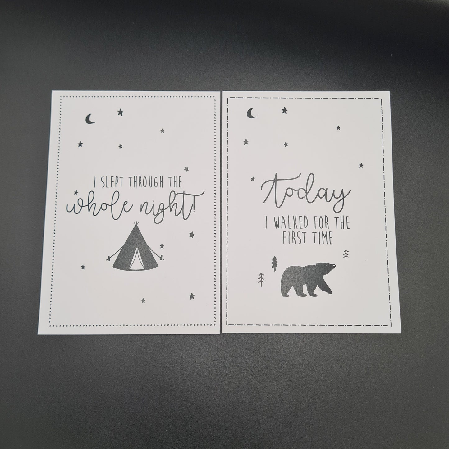 Two white baby milestone cards. The card on the left contains the text "I slept through the whole night" and an illustration of a tent with stars and the moon. The card on the right contains an illustration of stars, the moon and a bear and features the text "Today I walked for the first time".