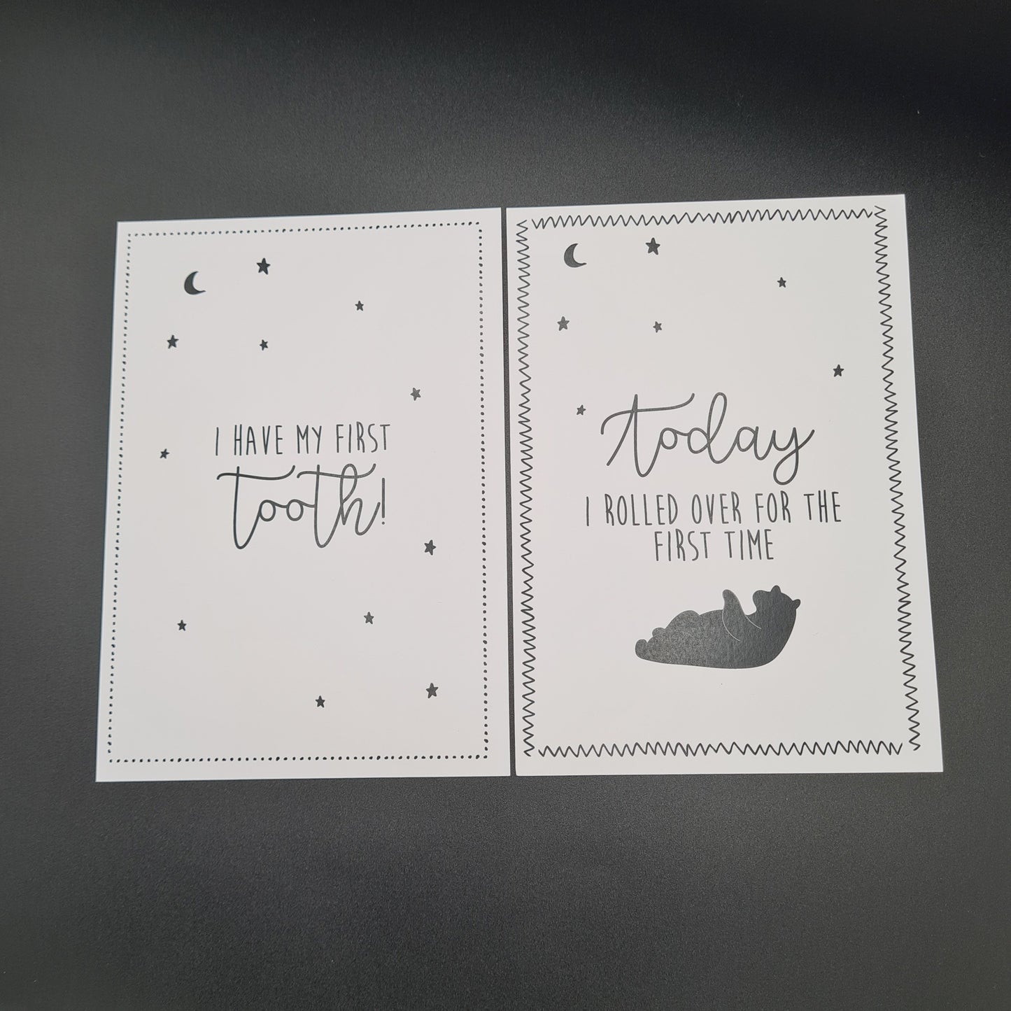 Two white baby milestone cards. The card on the left features the text "I have my first tooth" and illustrations of stars and the moon. The card on the right features illustrations of the moon, stars at the top and a bear that is rolling at the bottom. The text in the middle says "Today I rolled over for the first time".