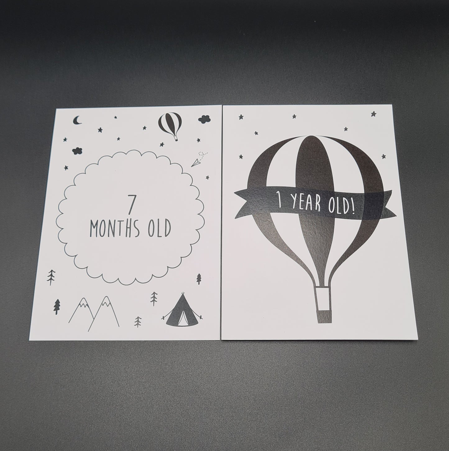 Two baby milestone cards, white in colour. The card on the left has illustrations of stars, the moon and a balloon at the top, the text "7 months old" in the middle and illustrations of mountains, trees and a tent at the bottom. The card on the right contains a large illustration of a balloon, stars above it and the text "1 Year Old" on a banner across the balloon.