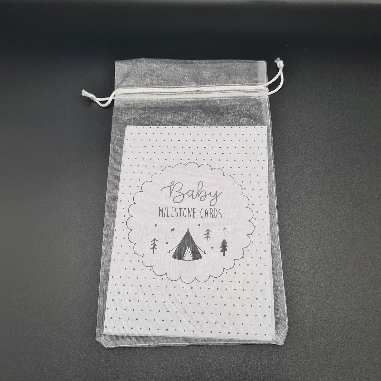 Pack of white Baby Milestone cards encased in a white organza bag. The visible top card is covered with black dots and has the text "Baby Milestone Cards" written inside a circle in the middle with an illustration of a tent and some trees.