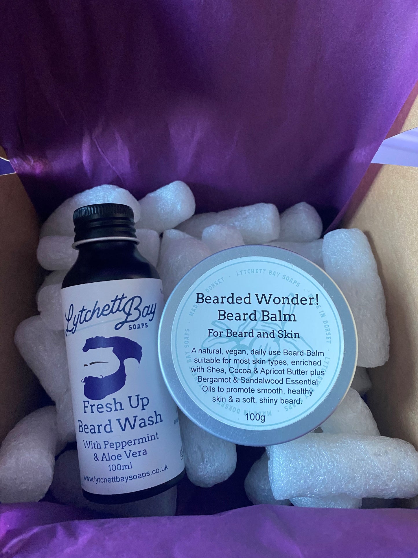 Gift box lined with purple tissue paper and containing white packing peanuts. Contents of the gift box are a "Fresh Up Beard Wash" bottle and "Bearded Wonder! Beard Balm" from Lytchett Bay Soaps.