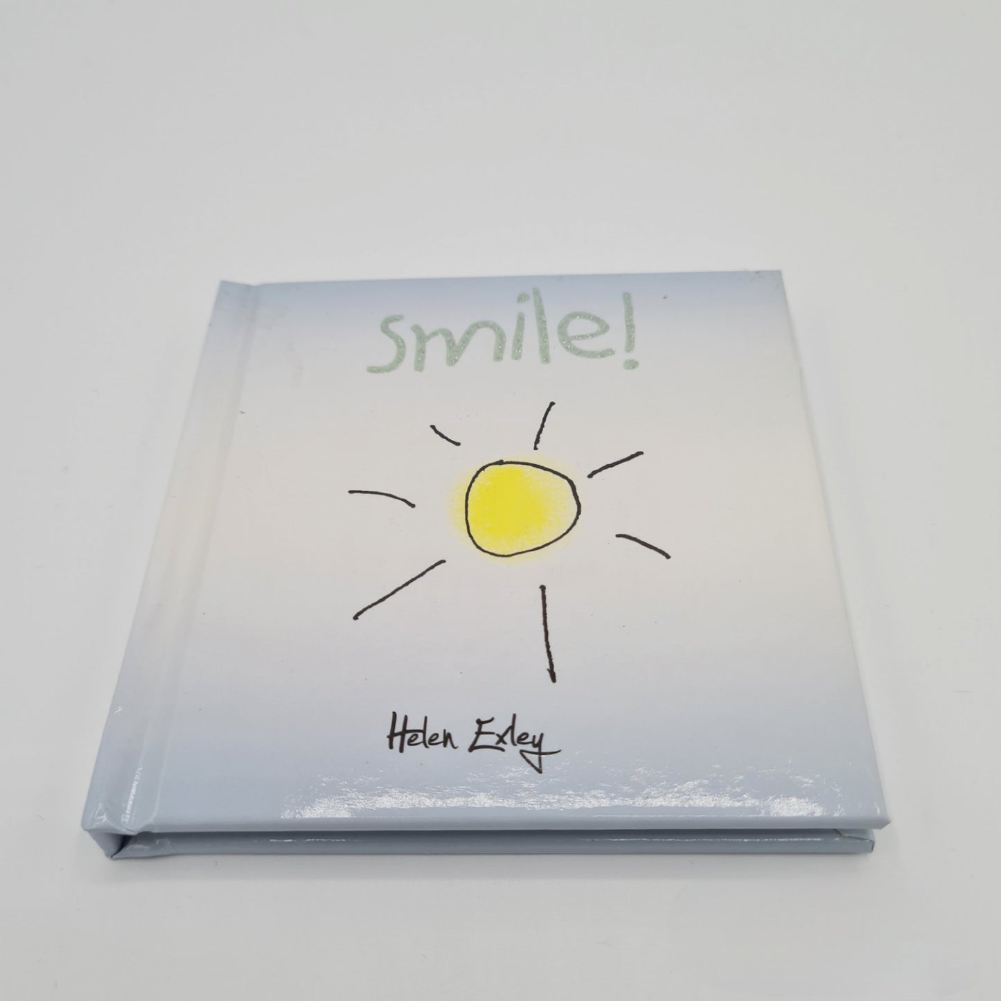 Front cover of little square book with the word "Smile" written in silver sparkly font at the top. The book is pale blue in colour and there is an illustration of a bright yellow sun in the middle of the page.