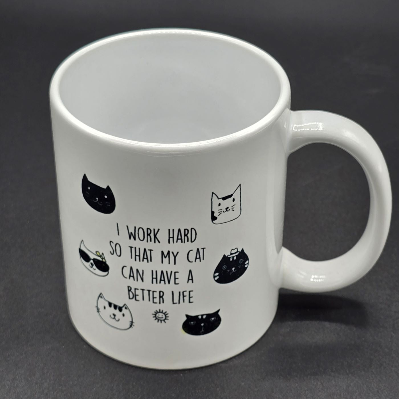 White mug with handle. Design on mug features several illustrations of cat faces and the text "I work hard so that my cat can have a better life".
