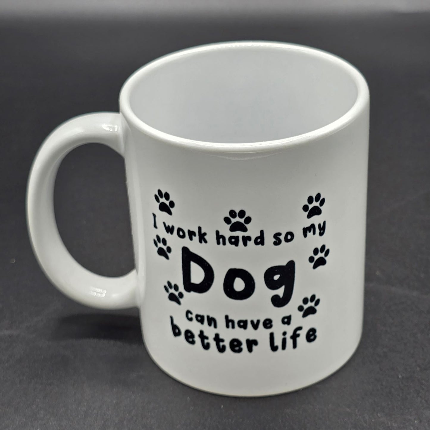 White mug with handle. Black text, surrounded by several black pawprints, says "I work hard so my Dog have can have a better life".