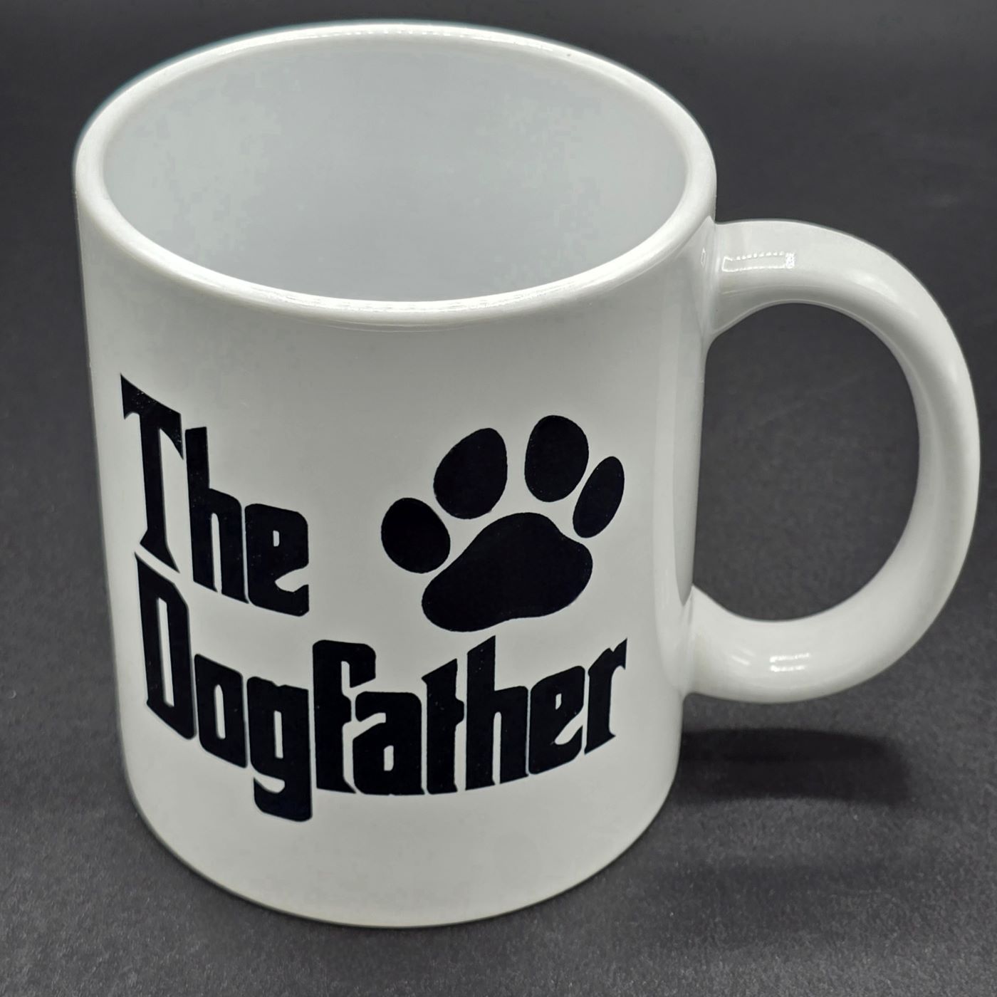 Plain white mug with white handle. Black text on the mug says "The Dogfather" and there is a paw print next to the text.