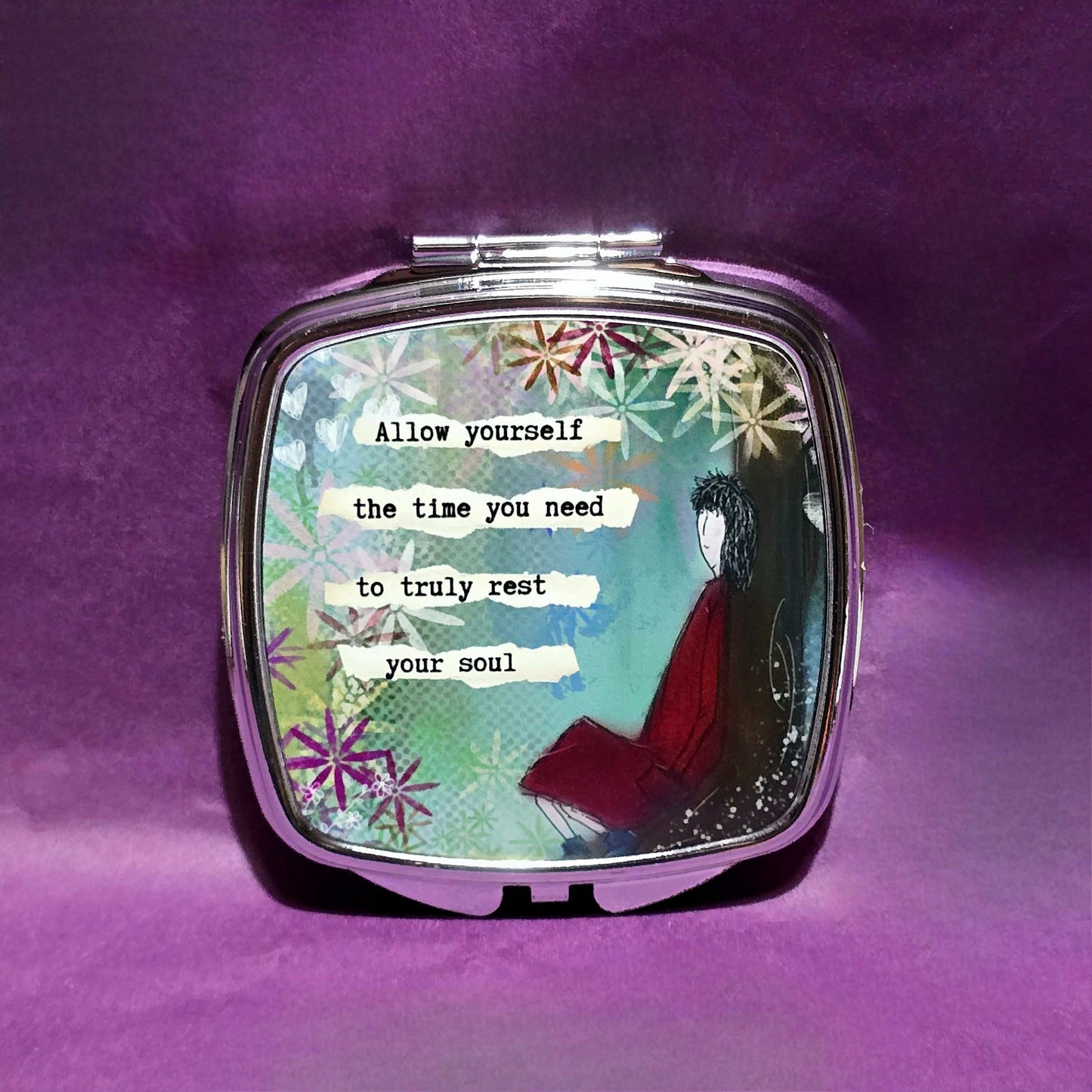 Silver compact mirror. The lid of the mirror has a sketch of a lady dressed in red sitting against a tree and features the text "Allow yourself the time you need to truly rest your soul".