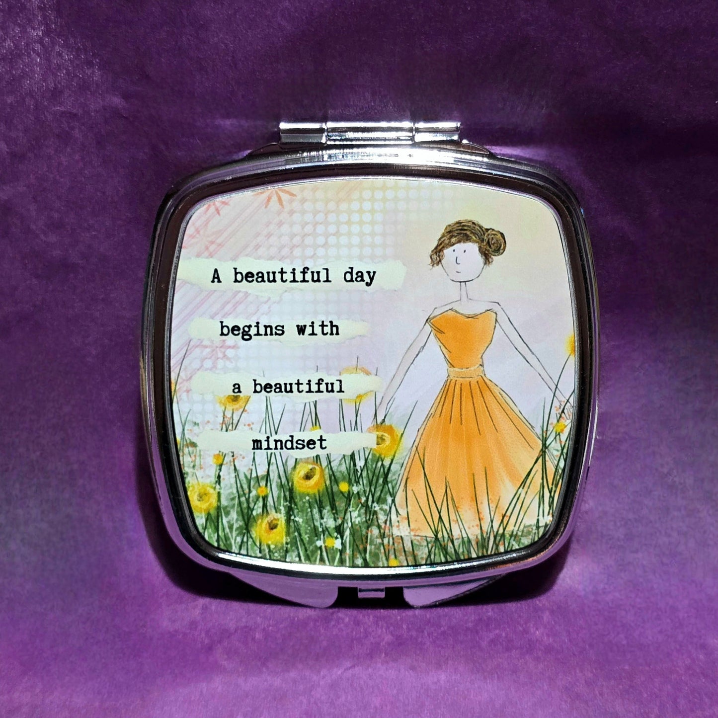 Square silver compact mirror. The lid features a sketch of a lady in a yellow dress standing in a field of yellow flowers. To the left of the lady features the text "A beautiful day begins with a beautiful mindset".