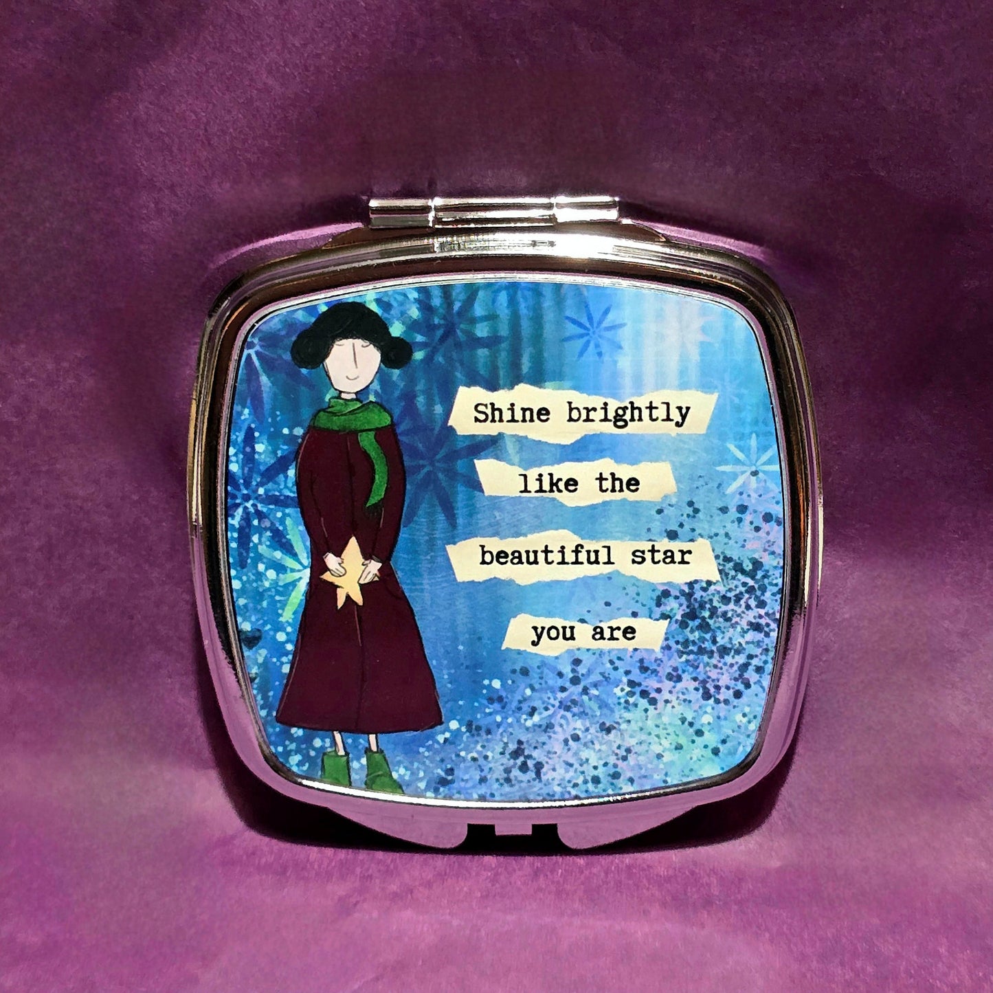 Square silver compact mirror. On the lid there is a sketch of a lady wearing a burgundy coat and green scarf and shoes. To the right of the lady features the text "Shine brightly like the beautiful star you are".