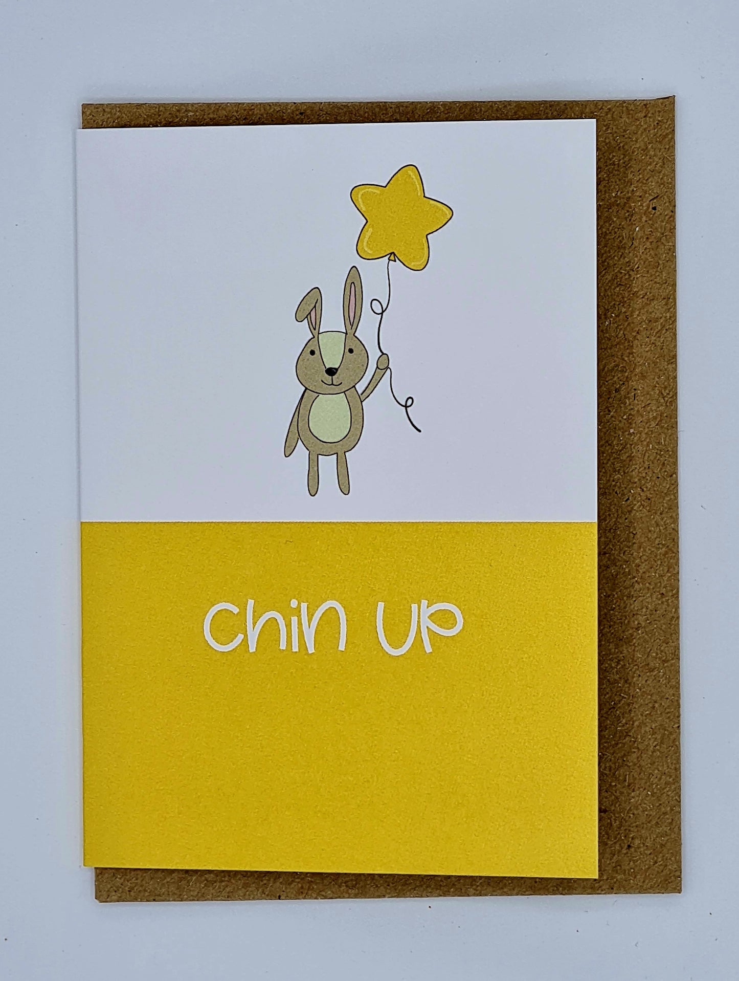 Mini A7 card with brown envelope behind. The top half of the card is white with a drawing of a rabbit holding a yellow star shaped balloon whilst the bottom half of the card is yellow with the words "Chin Up" written in white.