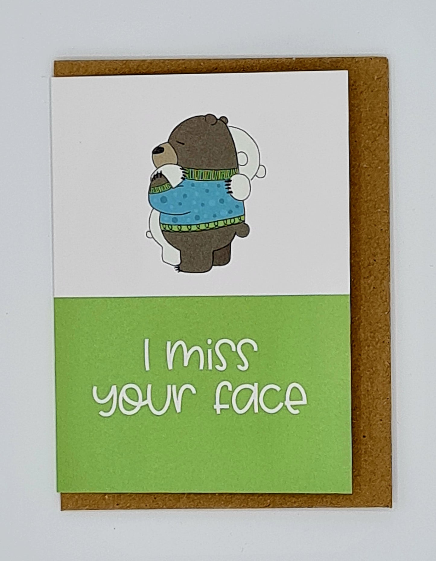 A7 mini card with brown envelope behind. The top half of the card is white and features a drawing of a brown bear wearing a blue jumper hugging a white bear. The bottom half of the card is green with the words " I Miss Your Face" written in white.