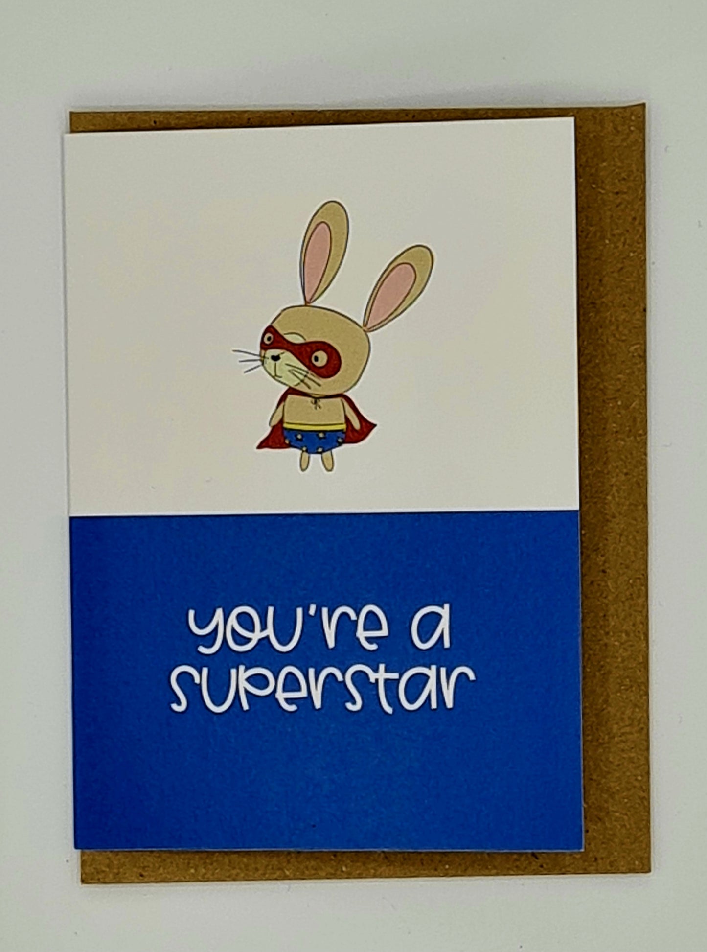 A7 mini card with brown envelope. The top half of the card is white with a drawing of a rabbit dressed as a superhero whilst the bottom half of the card is blue with the words "You're a Superstar" written in white.