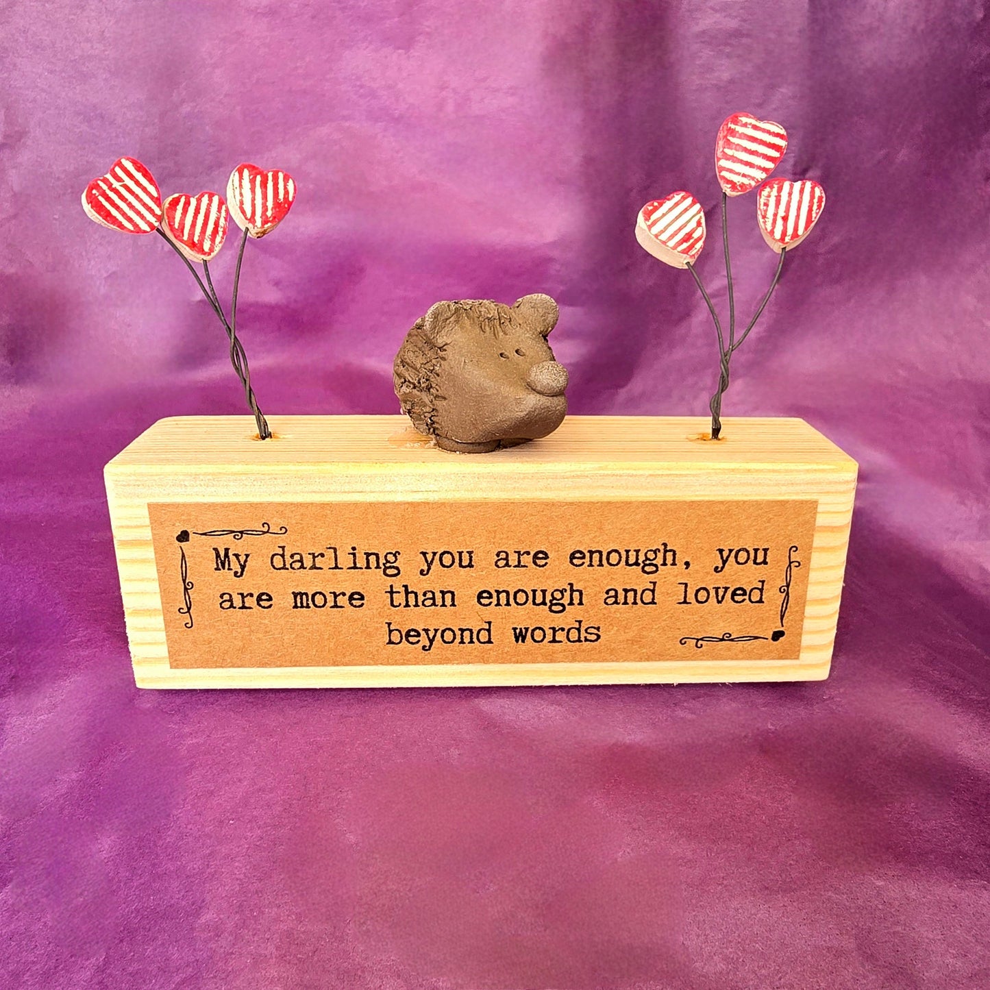 Mini sculpture made from a wooden block with a ceramic hedgehog sitting on top. To each side of the hedgehog there are three red and white striped hearts attached to wire. On the front of the wooden block there is a brown label with the text "My darling you are enough, you are more than enough and loved beyond words."