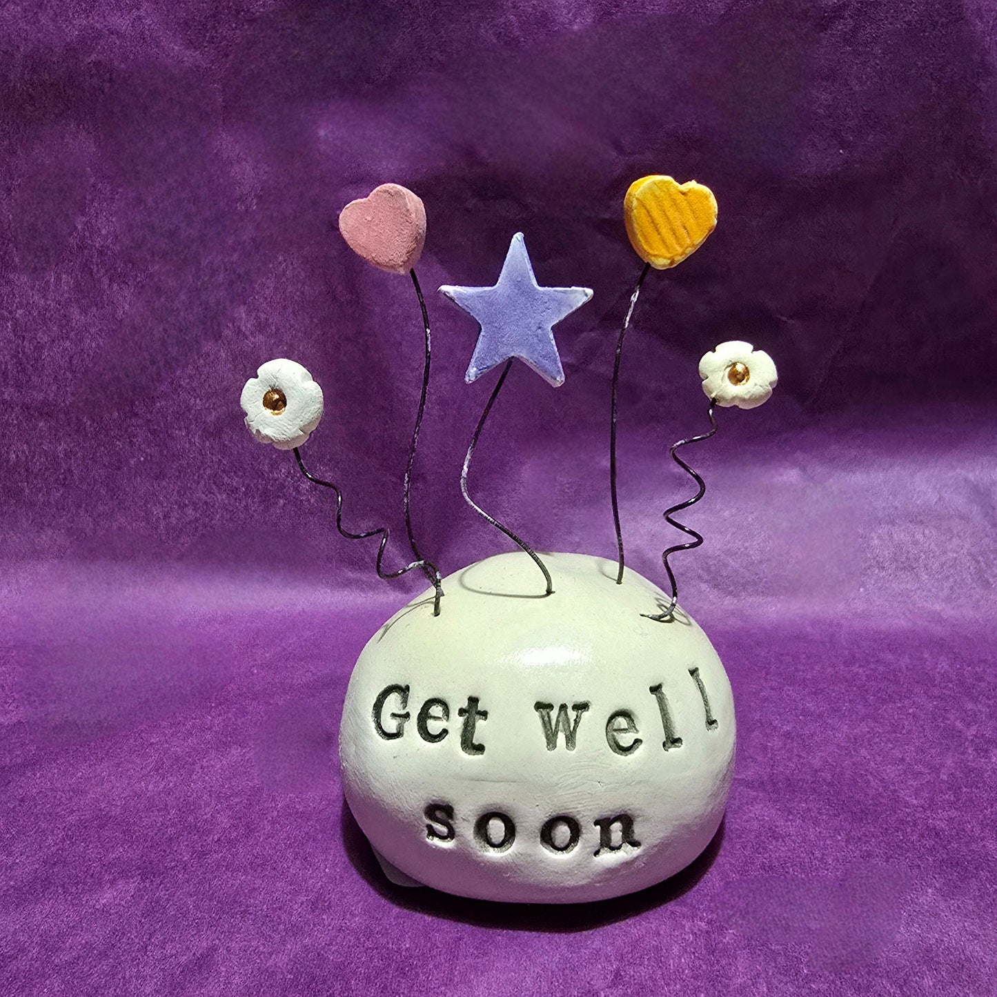 White ceramic dome with the words "Get Well Soon" embossed onto it. The top of the dome features five wires sprouting out, from left to right the wires feature a white flower, pink heart, blue star, yellow heart and white flower.