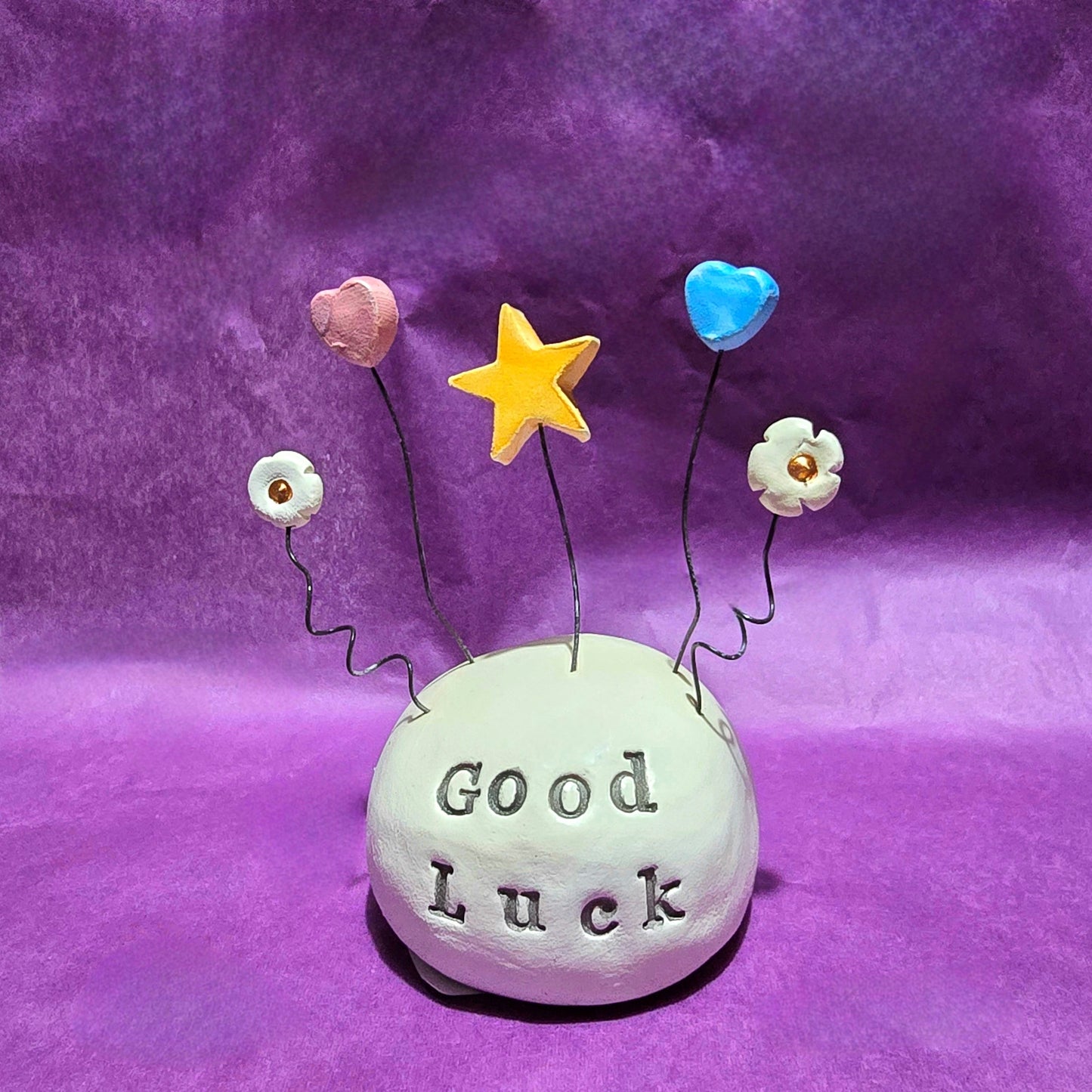 White ceramic dome with the words "Good Luck" embossed onto it. The top of the dome features five wires sprouting out, from left to right the wires feature a white flower, pink heart, yellow star, blue heart and white flower.