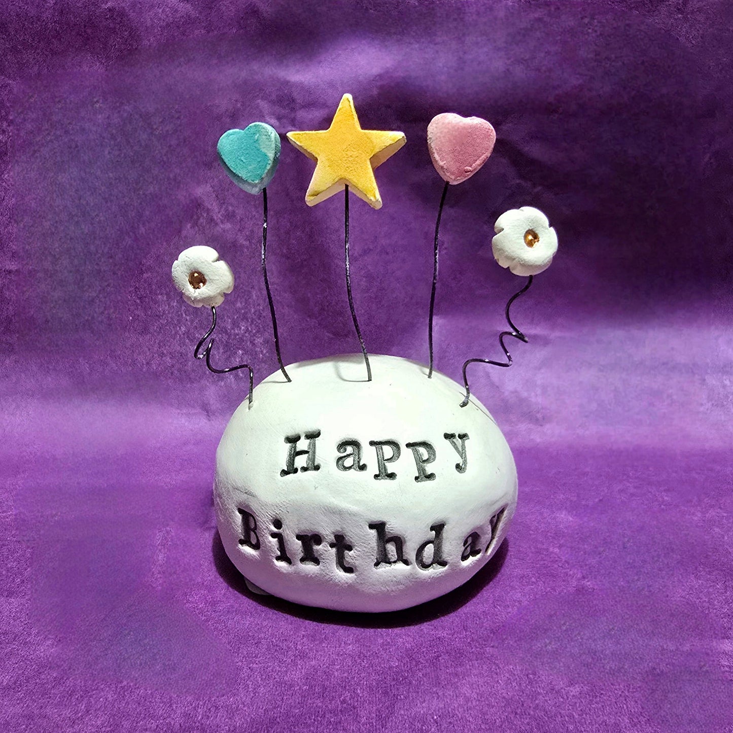 White ceramic dome with the words "Happy Birthday" embossed onto it. The top of the dome features five wires sprouting out, from left to right the wires feature a white flower, blue heart, yellow star, pink heart and white flower.