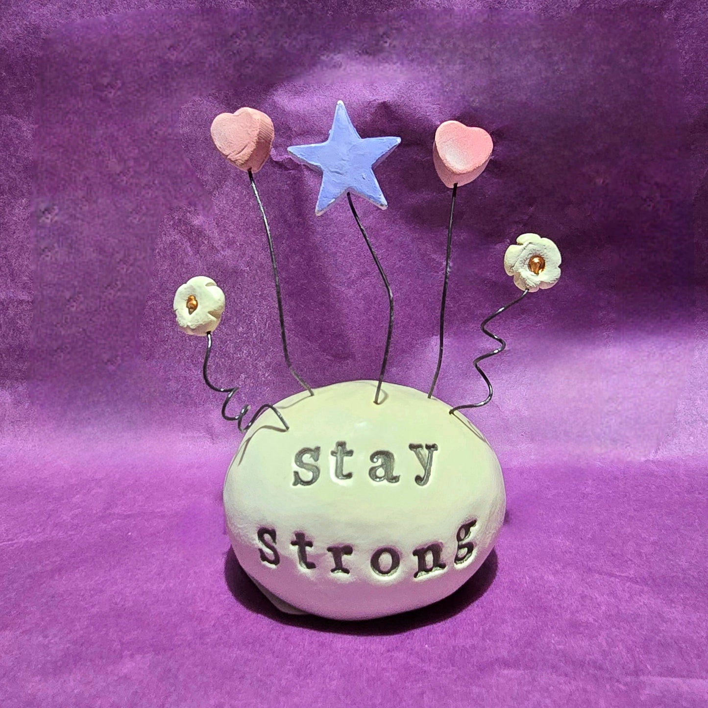White ceramic dome with the words "Stay Strong" embossed onto it. The top of the dome features five wires sprouting out, from left to right the wires feature a white flower, pink heart, blue star, pink heart and white flower.