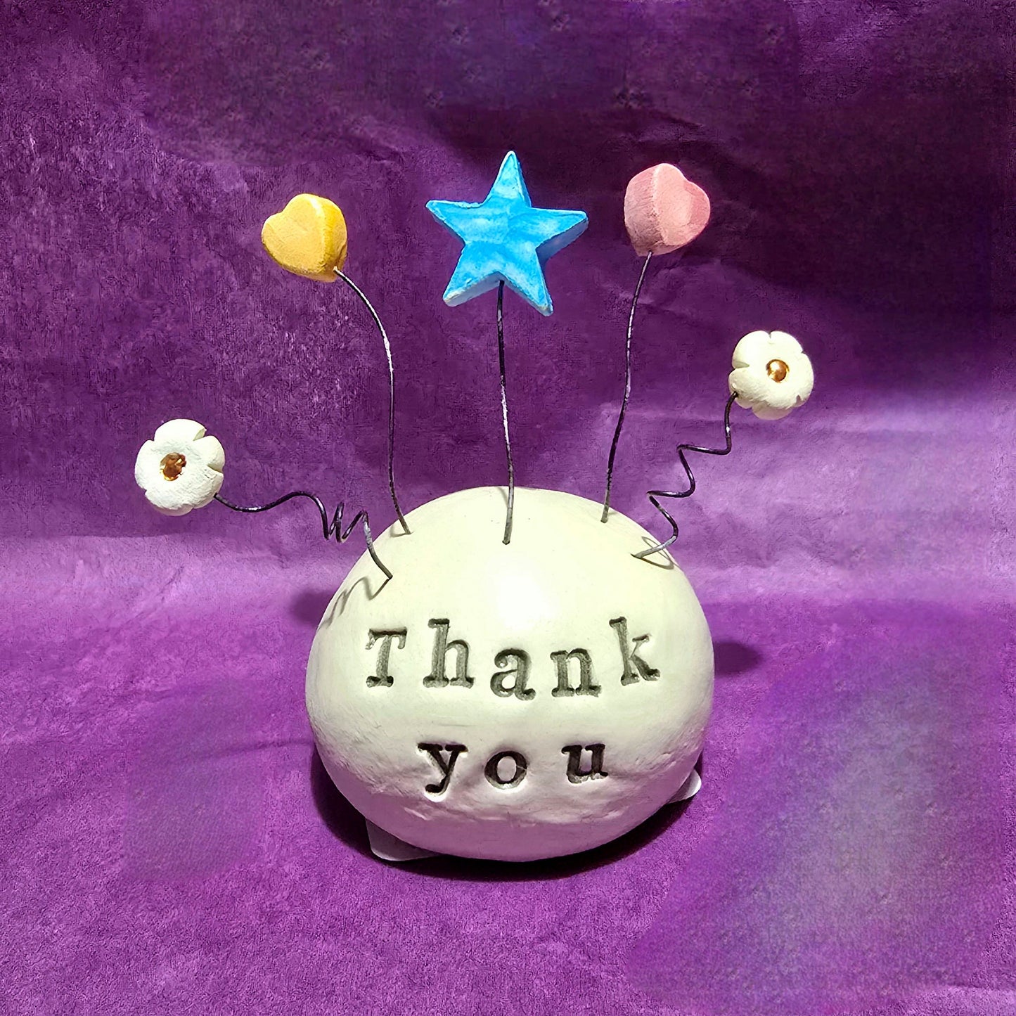 White ceramic dome with the words "Thank You" embossed onto it. The top of the dome features five wires sprouting out, from left to right the wires feature a white flower, yellow heart, blue star, pink heart and white flower.