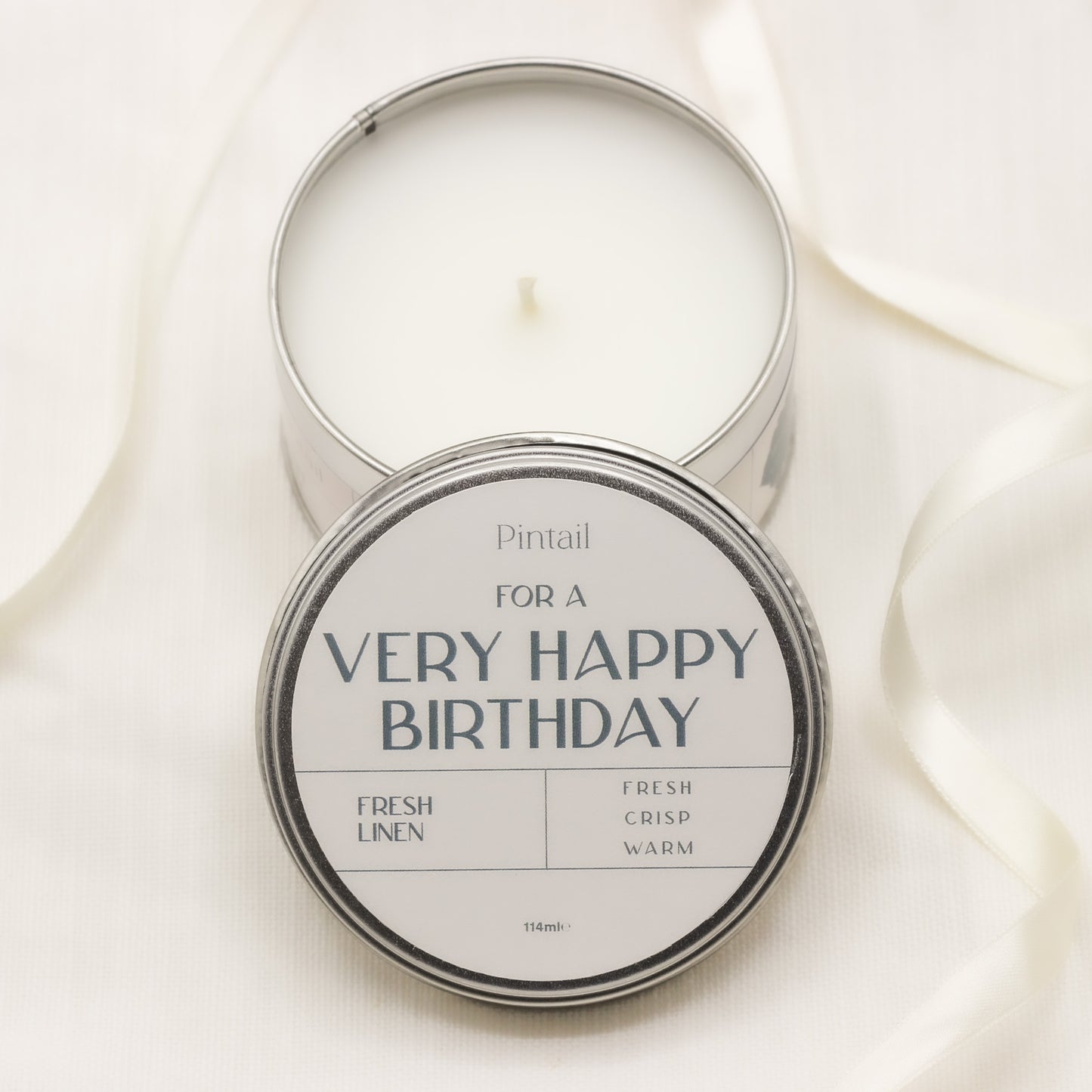 Open candle tin with the bottom half showing a white candle and wick, overlaid by the lid of the candle tin from Pintail with a white label containing the text "For a Very Happy Birthday", "Fresh Linen", "Fresh, Crisp, Warm".