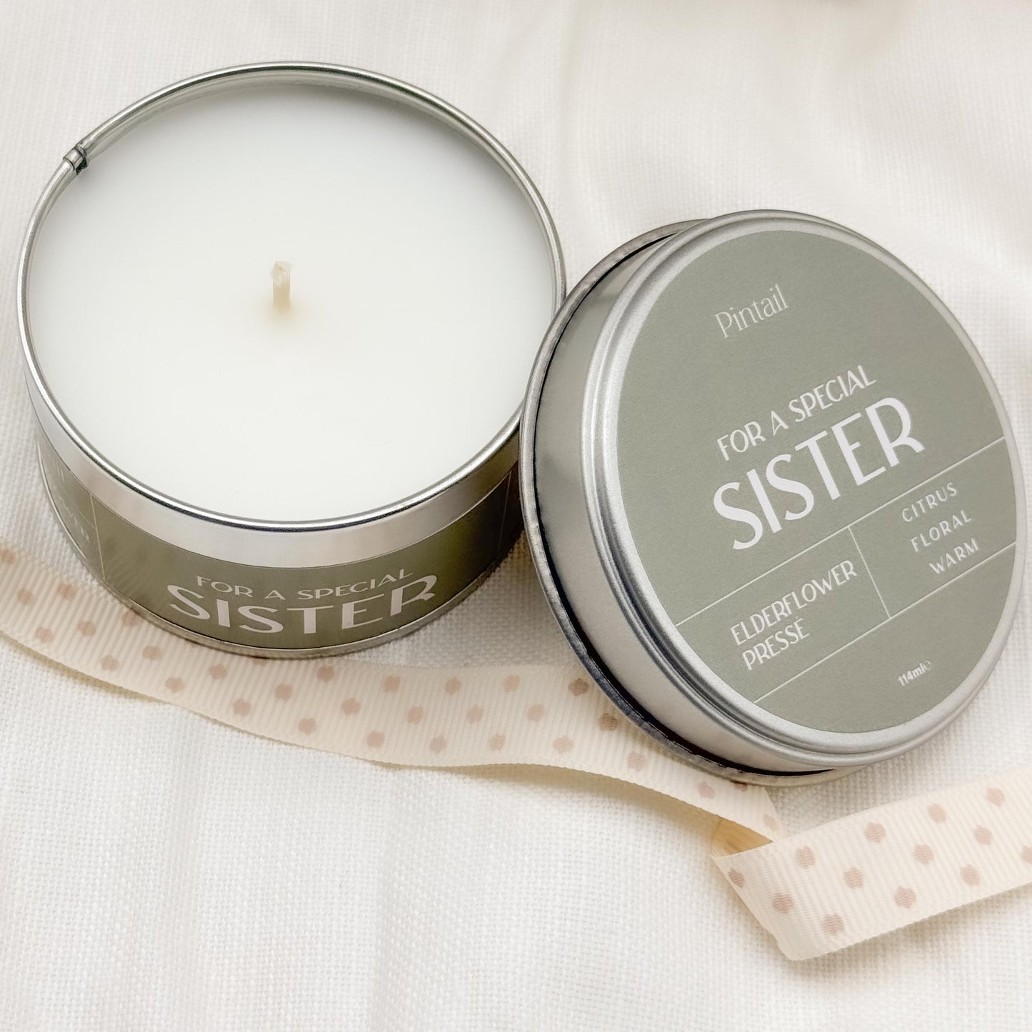 Open candle tin showing the white candle and wick on the left hand side of the image. On the right hand side of the image, the candle lid is leaning against the tin and contains the wording "For a Special Sister" "Elderflower Presse", "Citrus, Floral, Warm".
