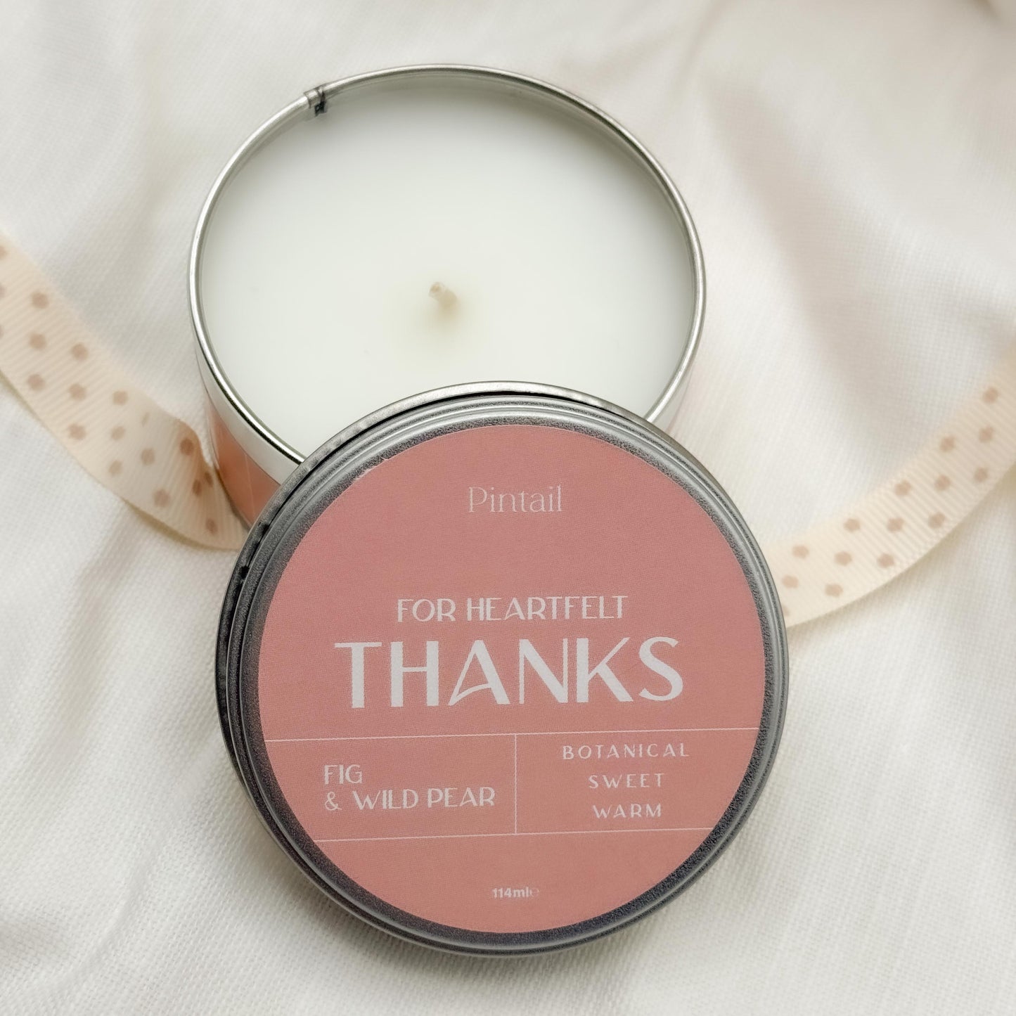 Open candle tin with the bottom half showing a white candle and wick, overlaid by the lid of the candle tin from Pintail with a pink label and the wording "For Heartfelt Thanks", "Fig & Wild Pear", "Botanical, Sweet, Warm".