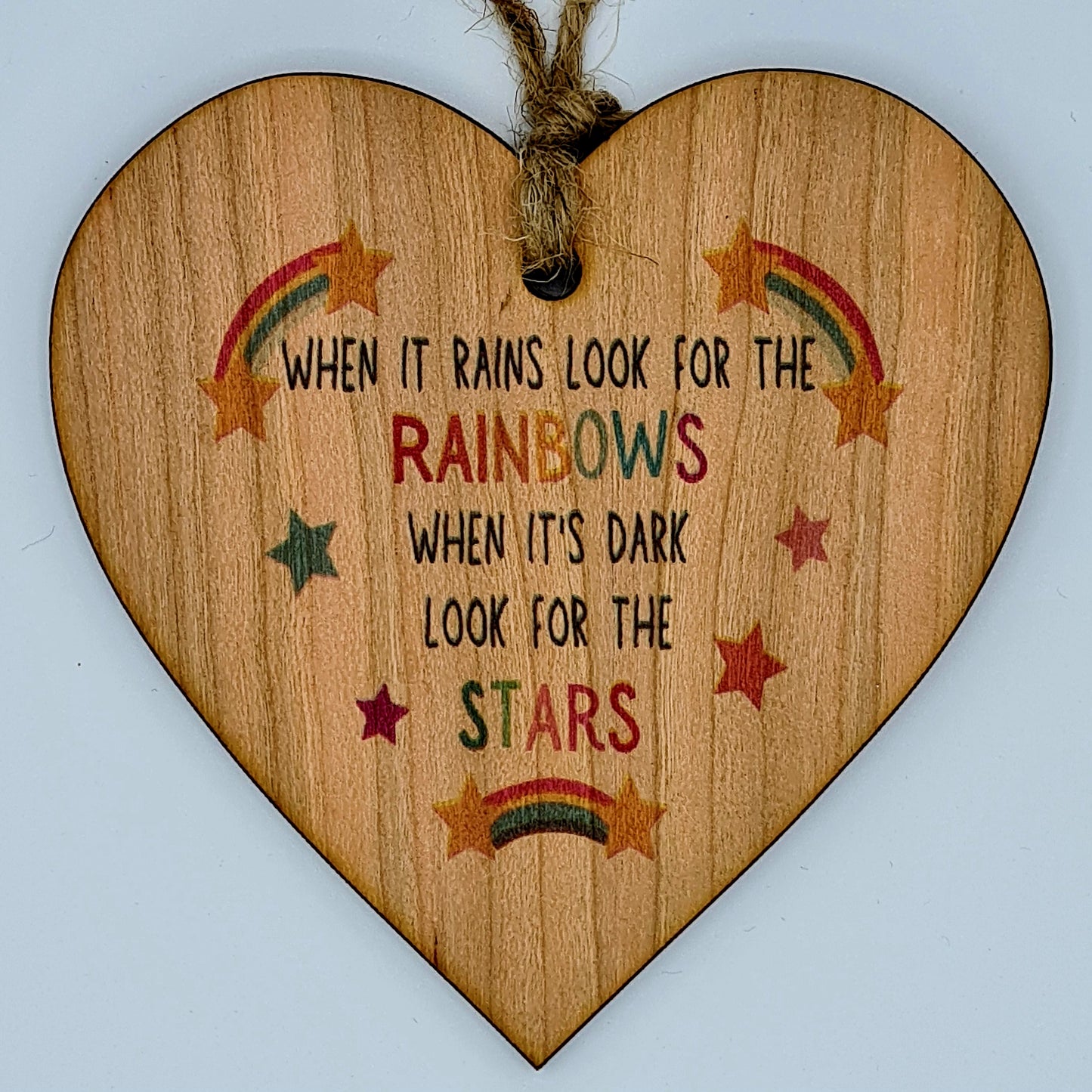 Heart shaped wooden plaque with the slogan "When it rains look for the rainbows, when it's dark look for the stars". There are little rainbows and stars drawn around the writing. At the top of the plaque, there is a jute string for hanging.