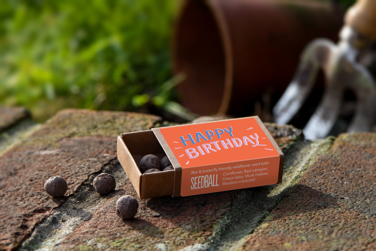 Brown match box sized box with orange label saying "Happy Birthday" on top. On the side of the box the text says "Bee & butterfly friendly wildflower seedballs: Cornflower; Red Campion; Oxeye Daisy; Musk Mallow; Meadow Cranesbill". Inside the box there are several brown seedballs and there are also three seedballs sitting next to the box.