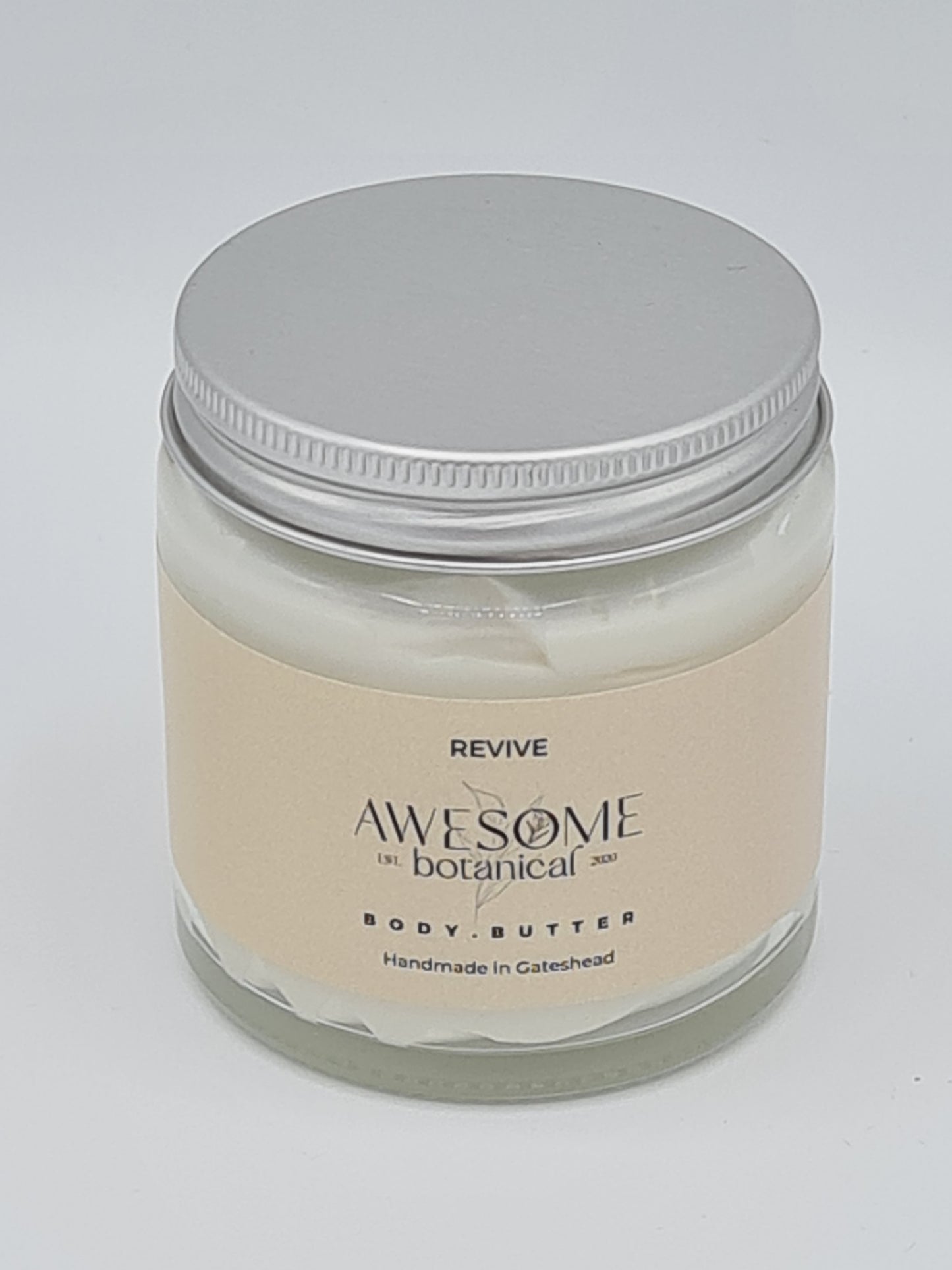 Glass jar of Body Butter with silver lid. Jar has cream label from Awesome Botanical with the name Revive.