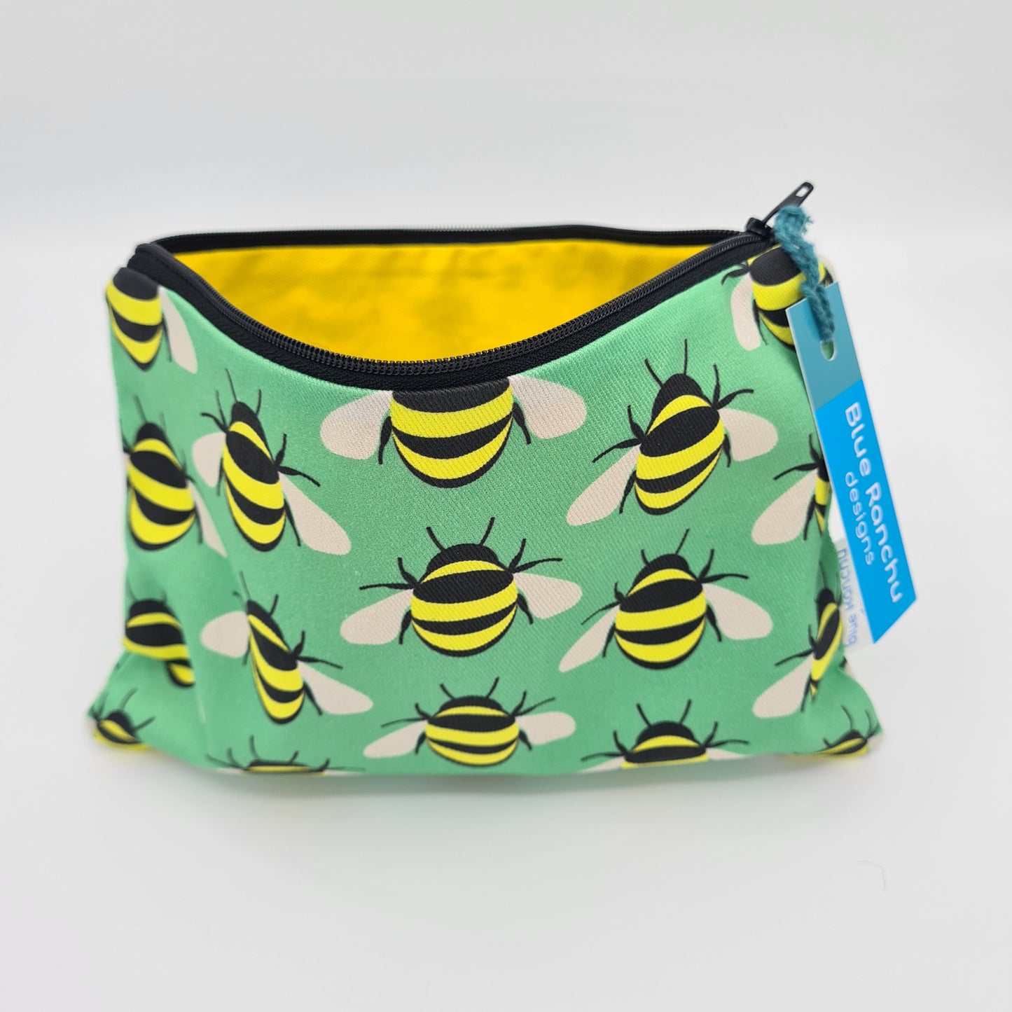 Green cotton accessories bag with print of yellow and black bees all over. The bag is open and unzipped showing the bright yellow cotton lining inside. There is a label for "Blue Ranchu Designs" attached to the zip.