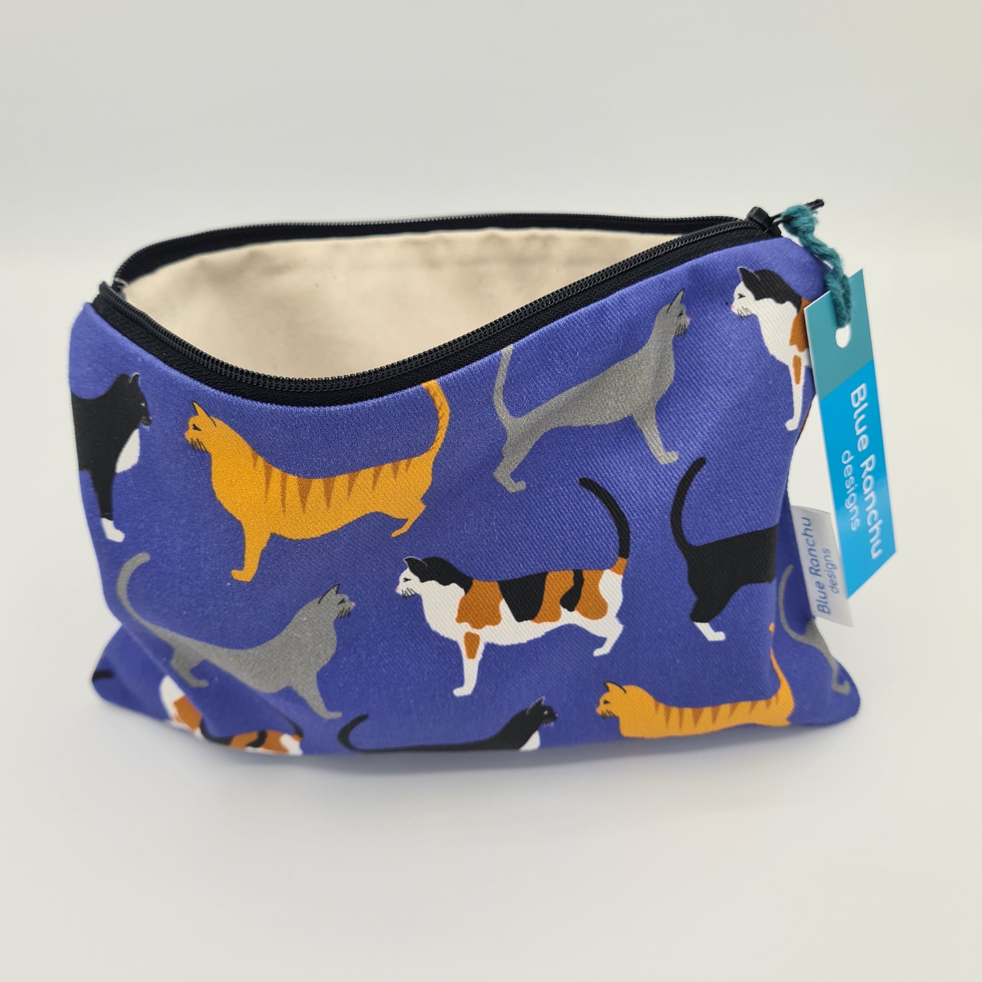 Blue cotton accessories bag with print of three cats all over. The bag is open and unzipped showing the cream cotton lining inside. There is a label for "Blue Ranchu Designs" attached to the zip.