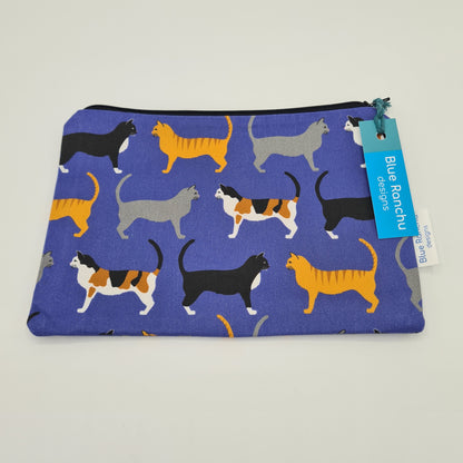 Blue cotton accessories bag with print of three cats all over. The first cat is black with a white chest, the second cat is ginger with dark stripes on its back and the third cat is grey. The bag features a black zip and there is a label attached saying "Blue Ranchu Designs".