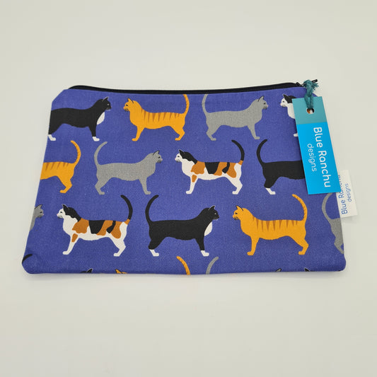 Blue cotton accessories bag with print of three cats all over. The first cat is black with a white chest, the second cat is ginger with dark stripes on its back and the third cat is grey. The bag features a black zip and there is a label attached saying "Blue Ranchu Designs".