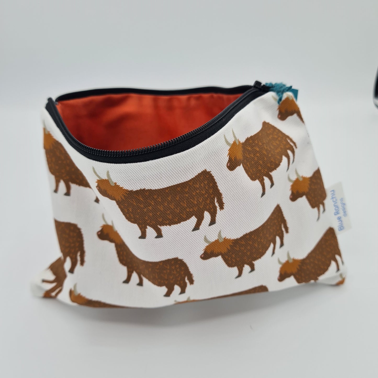 Cream cotton accessories bag with print of brown Highland Cows all over. The bag is open and unzipped showing the burnt orange coloured cotton lining inside. There is a label for "Blue Ranchu Designs" attached to the side of the bag.