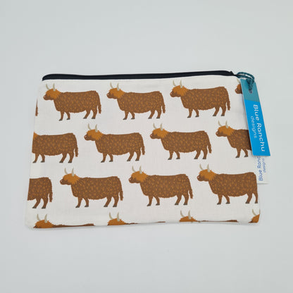 Cream cotton accessories bag with print of brown Highland Cows all over. The bag features a black zip and there is a label attached saying "Blue Ranchu Designs".