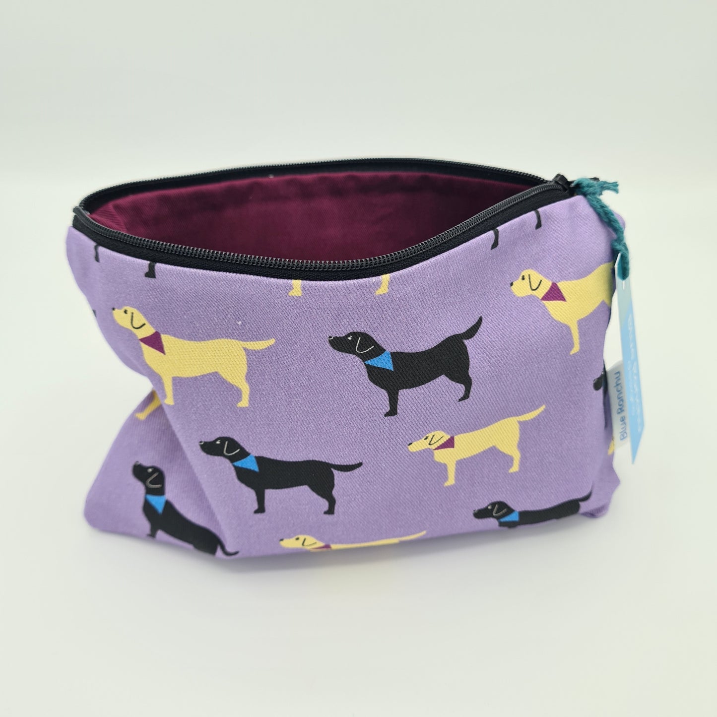 Lilac cotton accessories bag with print of black and yellow Labrador dogs all over. The bag is open and unzipped showing the burgundy coloured cotton lining inside. There is a label for "Blue Ranchu Designs" attached to side of the bag.