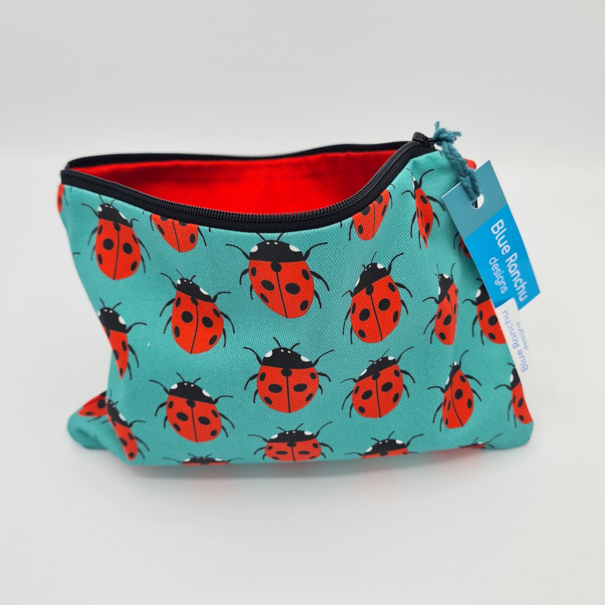 Turquoise cotton accessories bag with print of black and red Ladybirds all over. The bag is open and unzipped showing the red cotton lining inside. There is a label for "Blue Ranchu Designs" attached to the zip.