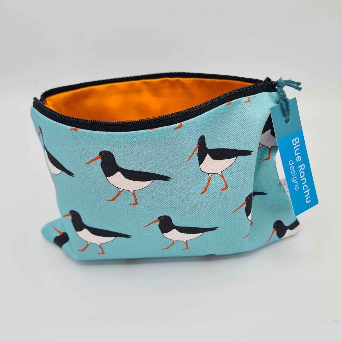 Light blue cotton accessories bag with print of black and white Oystercatcher birds all over. The bag is open and unzipped showing the bright orange cotton lining inside. There is a label for "Blue Ranchu Designs" attached to the zip.