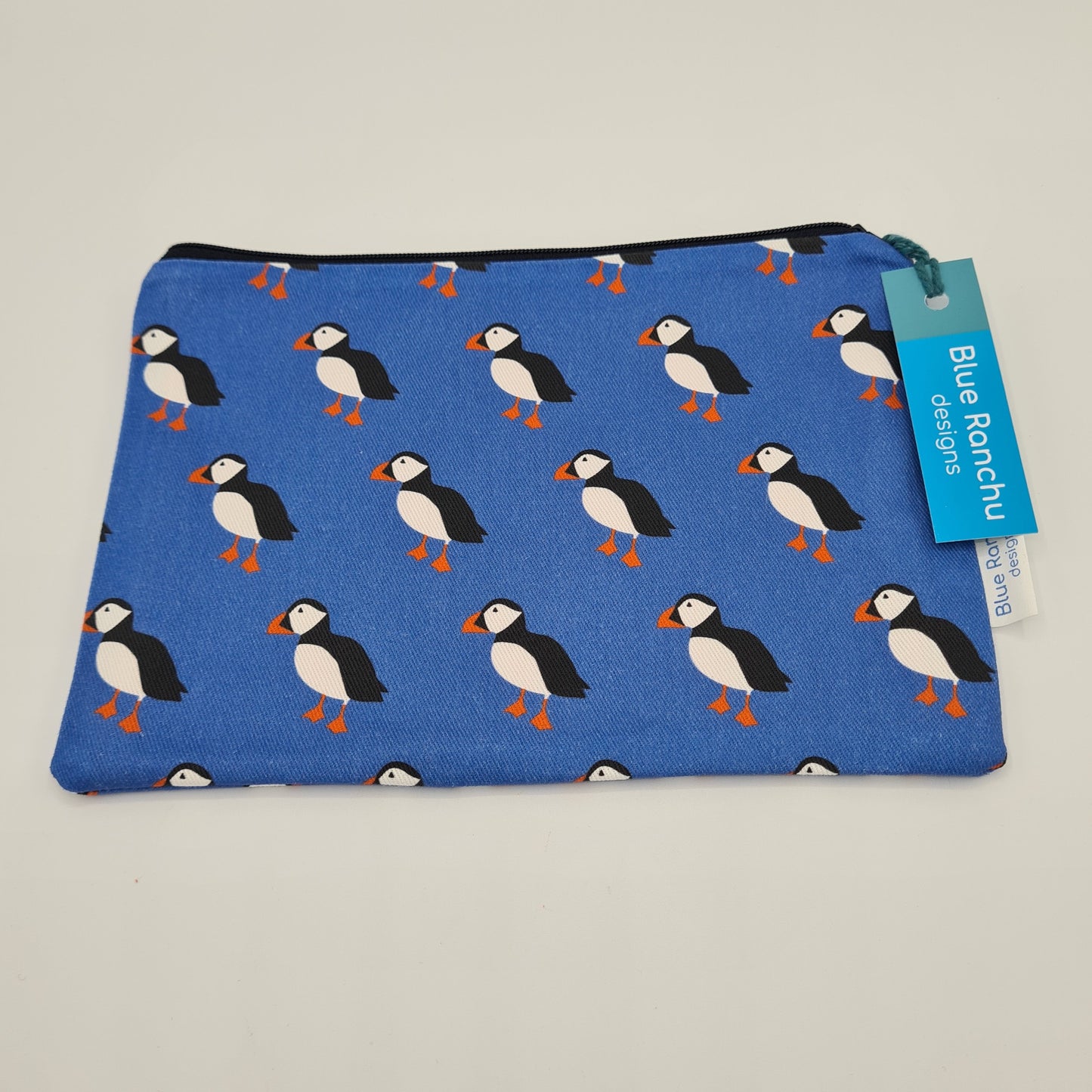 Blue cotton accessories bag with print of black and white Puffins with orange feet and beaks. The bag features a black zip and there is a label attached saying "Blue Ranchu Designs".