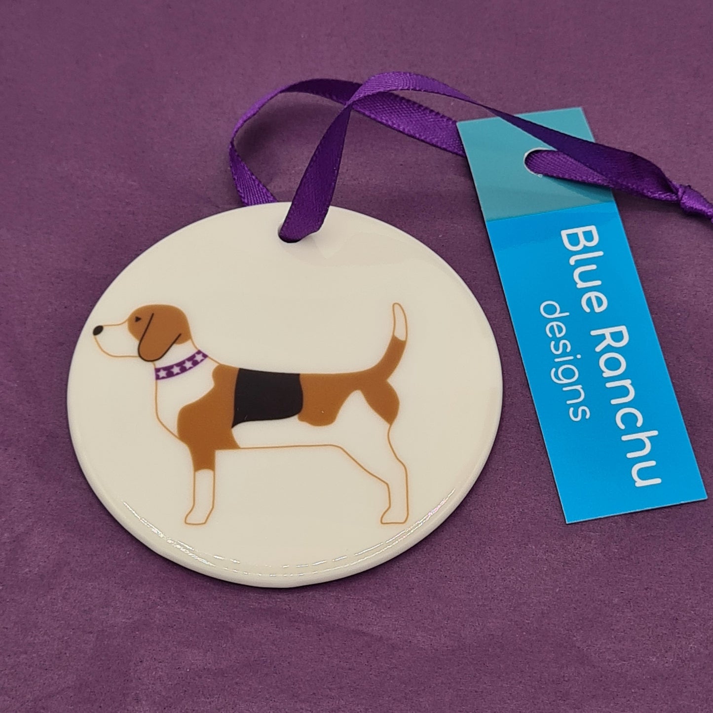 Circular white ceramic hanging decoration featuring a print of a brown, white and black Beagle with a purple collar around its neck. There is a purple ribbon attached for hanging and a label saying "Blue Ranchu Designs".