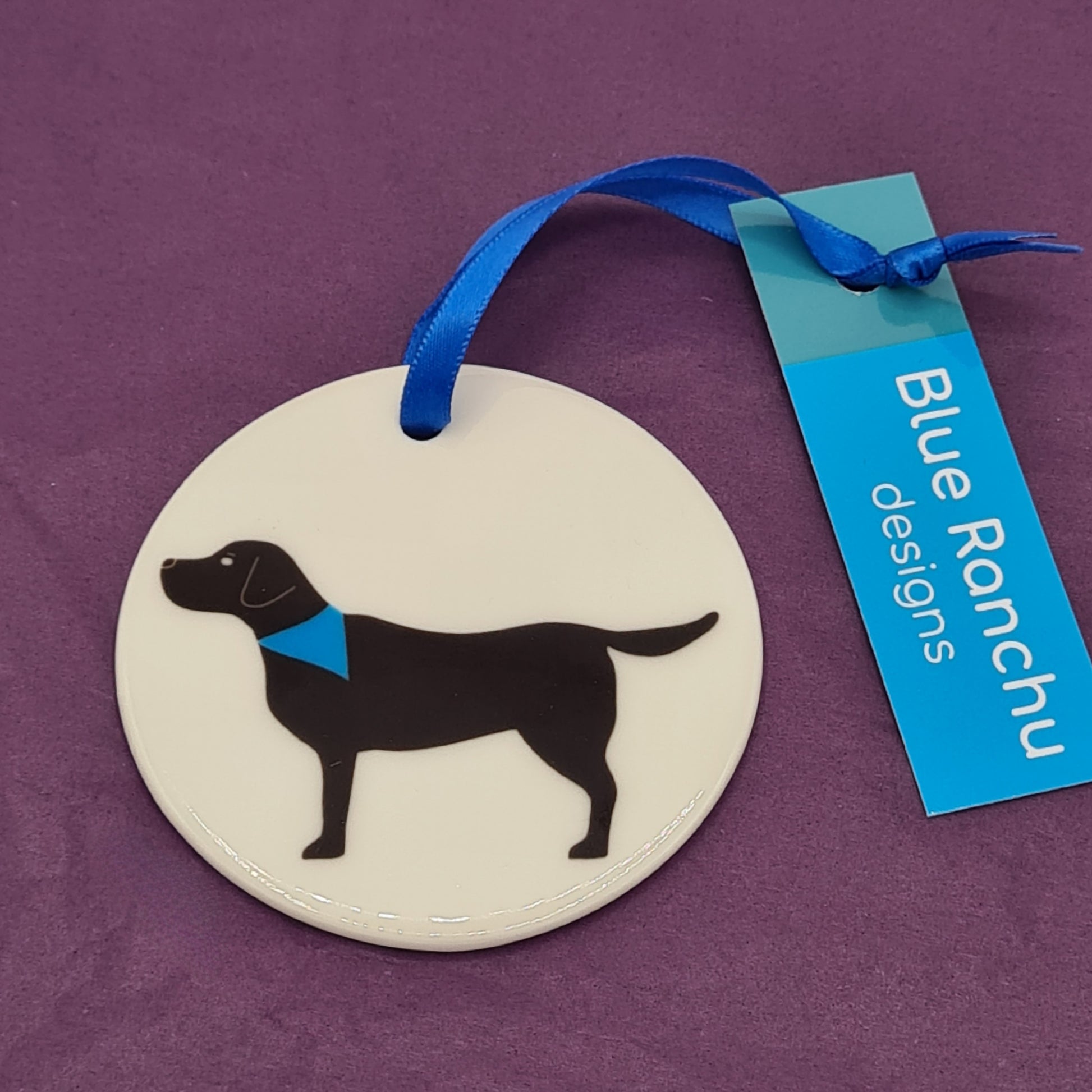 Circular white ceramic hanging decoration featuring a print of a black Labrador wearing a blue bandana around its neck. There is a blue ribbon attached for hanging and a label saying "Blue Ranchu Designs".