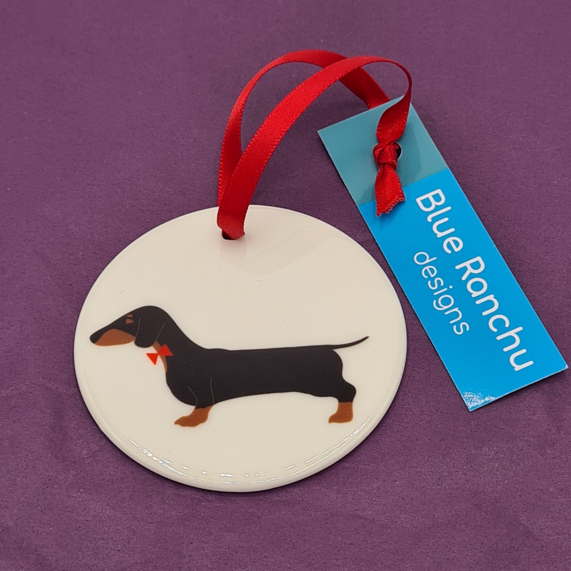 Circular white ceramic hanging decoration featuring a print of a black and tan Dachshund with a red bow tie on its neck. There is a red ribbon attached for hanging and a label saying "Blue Ranchu Designs".