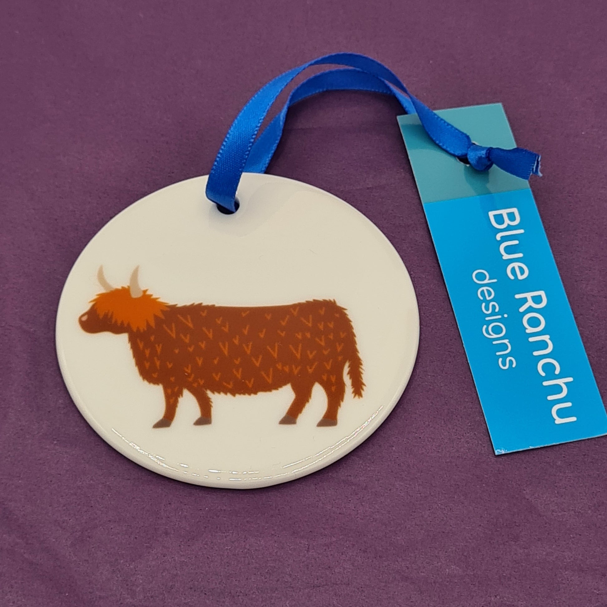 Circular white ceramic hanging decoration featuring a print of a Highland Cow. There is a blue ribbon attached for hanging and a label saying "Blue Ranchu Designs".