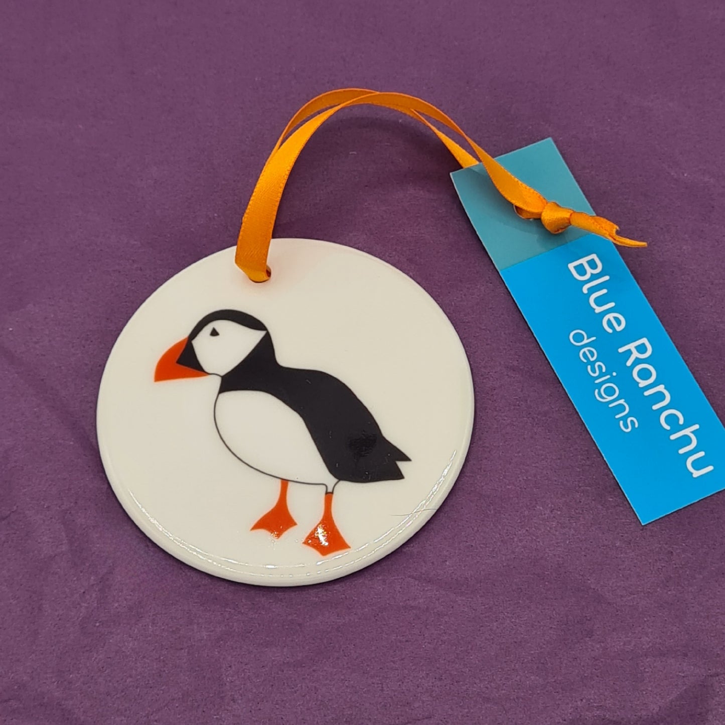 Circular white ceramic hanging decoration featuring a print of a black and white Puffin with orange beak and feet. There is an orange ribbon attached for hanging and a label saying "Blue Ranchu Designs".