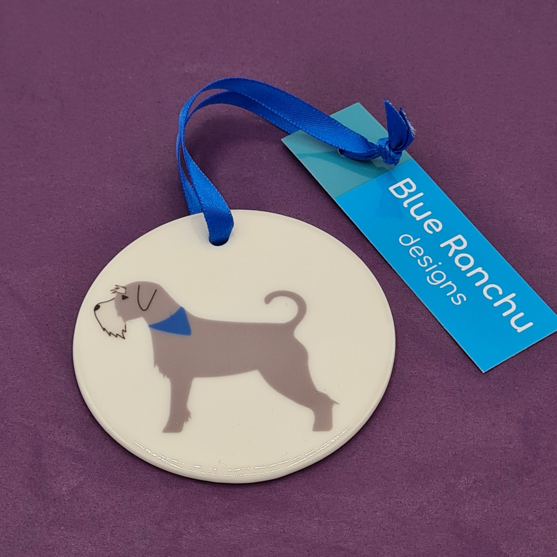 Circular white ceramic hanging decoration featuring a print of a grey Schnauzer wearing a blue bandana around its neck. There is a blue ribbon attached for hanging and a label saying "Blue Ranchu Designs".